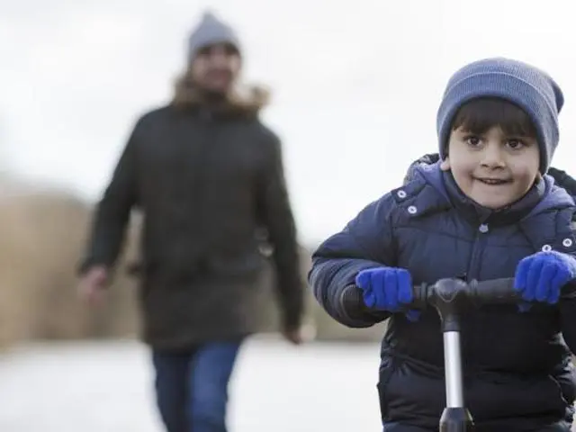 Boy scooting ahead of adult who is blurred out walking far behind.