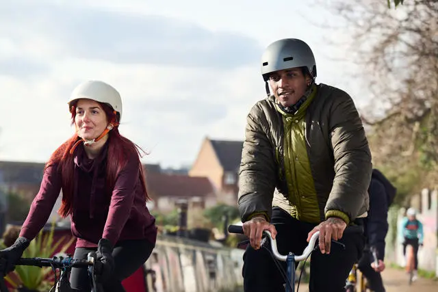 Two people cycling on a cycle path. One woman, one man. The woman is wearing purple, the man green. 