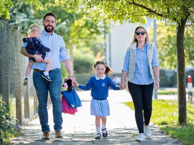 A couple with a toddler walk a small child to school on a sunny day