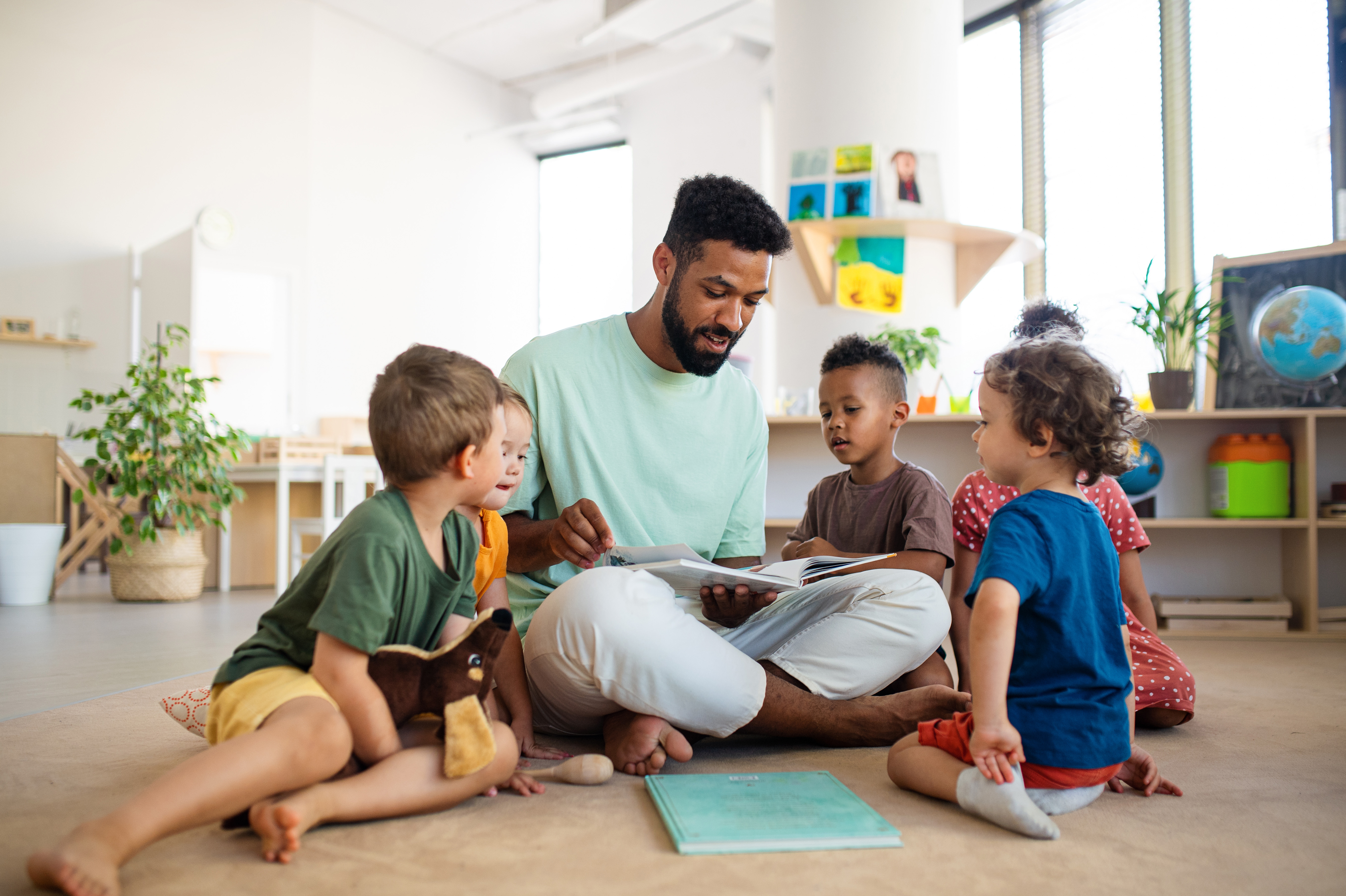 male adult sitting on the floor reading a book to a number of children in a nursery setting