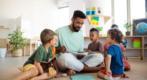 male adult sitting on the floor reading a book to a number of children in a nursery setting