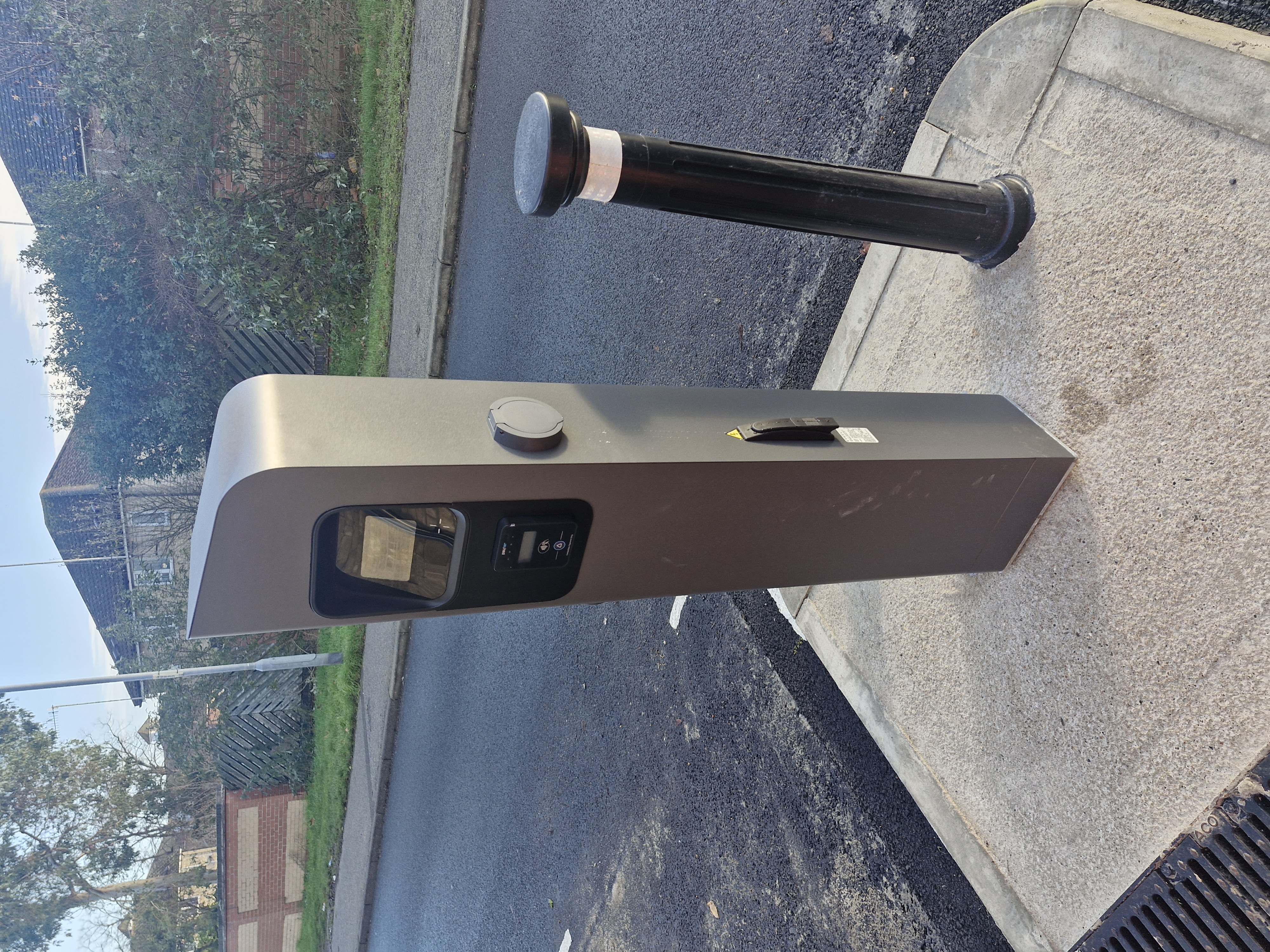 The chargepoint is grey and sleek looking. It is on a build-out on a tarmacked road, with a bollard to the right of it.