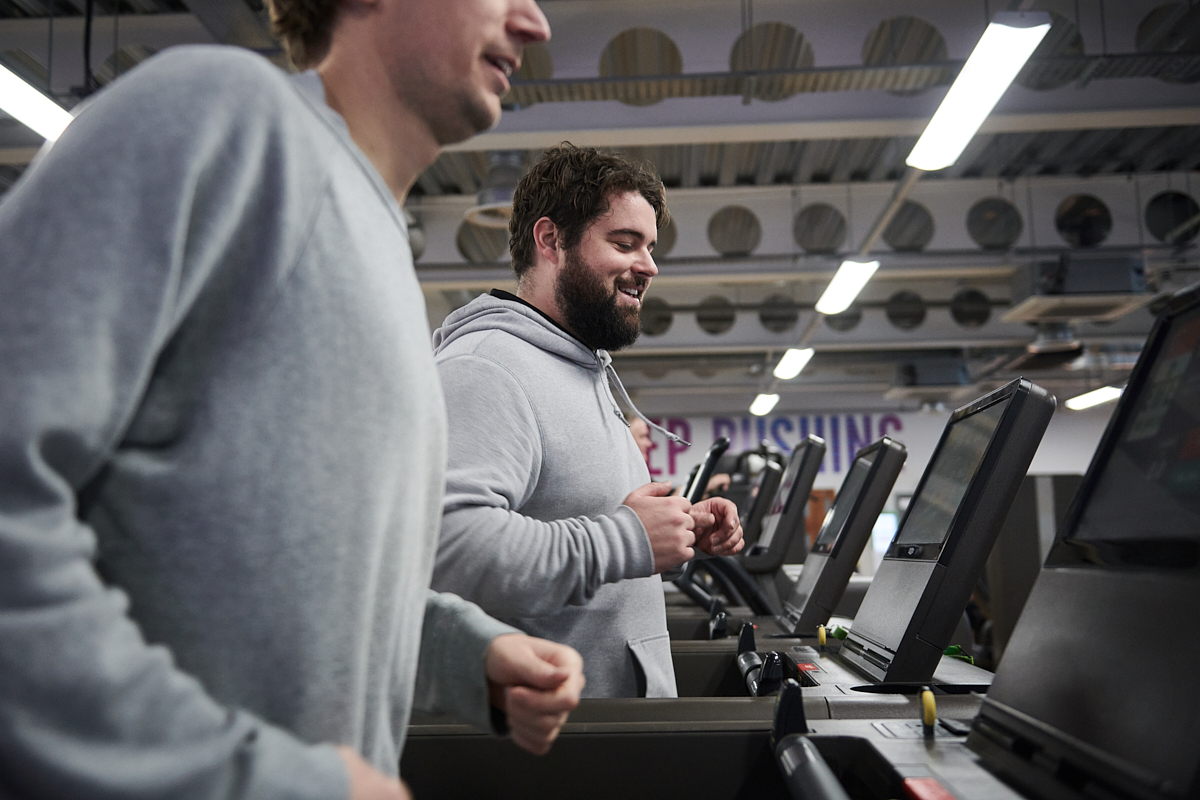 Two men in grey jumpers in the gym jogging on running machines