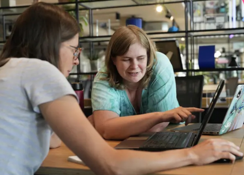 Two women are working on a shared laptop, one is pointing at the screen
