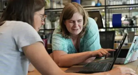 Two women are working on a shared laptop, one is pointing at the screen