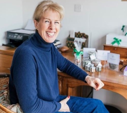 A lady sitting in a chair next to her desk. She is smiling. Her products are displayed on top of the desk.