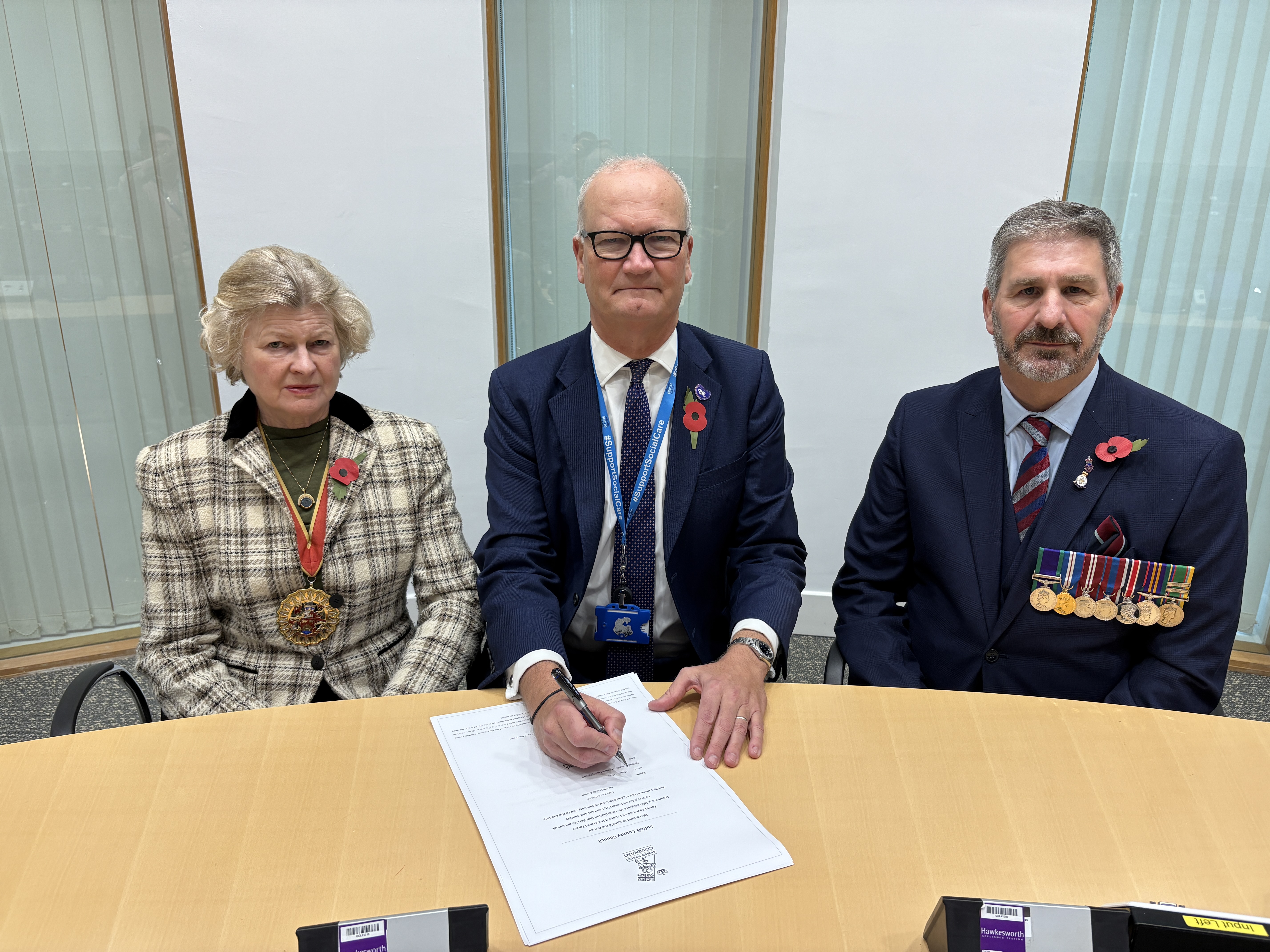 Three people smartly dressed, wearing medals and poppies, with the centre person signing