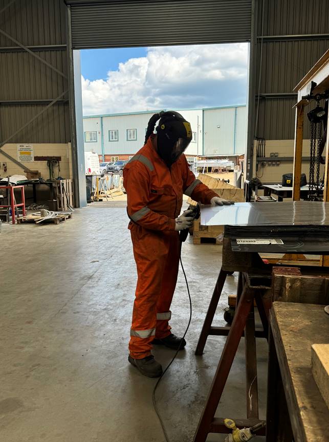 A person in protective orange clothing, working in a fabrication workshop