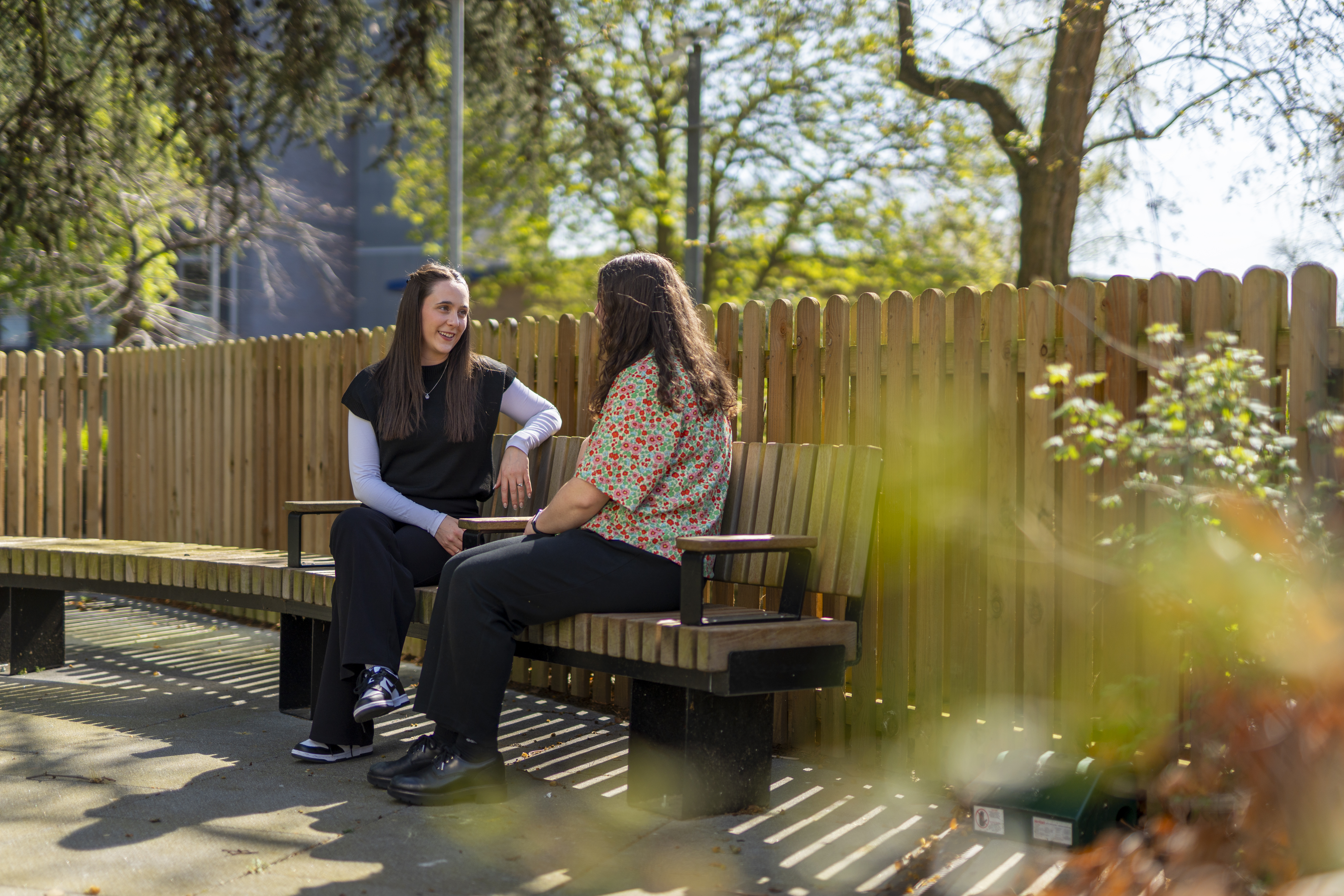 Two people sitting on a park bench looking at each other with a fence behind them. 