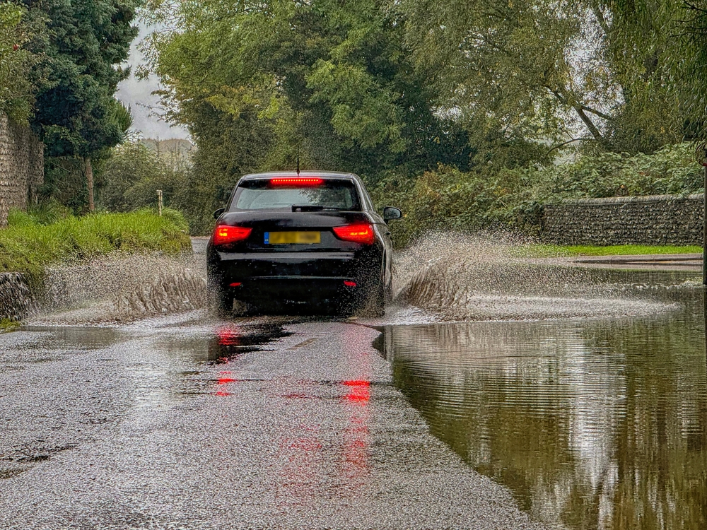 A black car drives through a flooded road, brown water splashes up from their tyers 