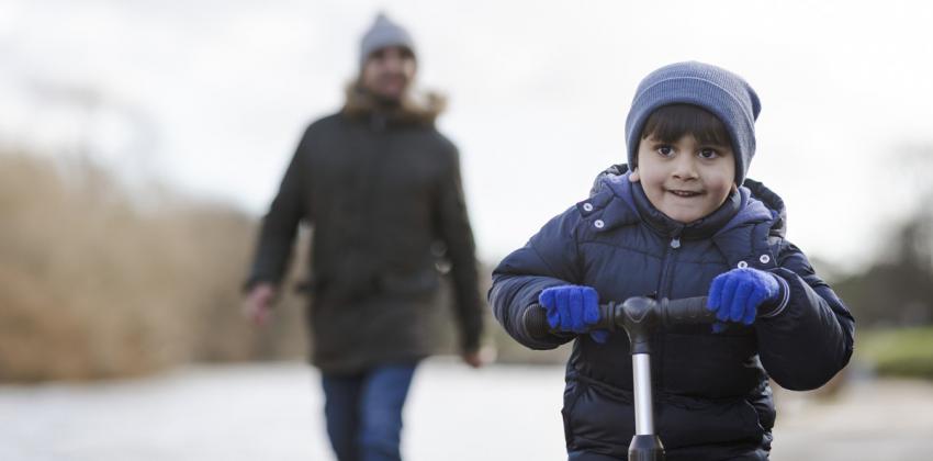Boy scooting ahead of adult who is blurred out walking far behind. 