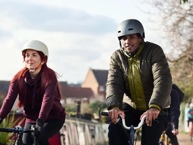 Two people cycling on a cycle path. One woman, one man. The woman is wearing purple, the man green. 