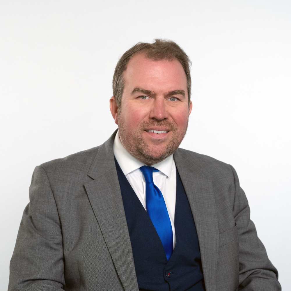Councillor Darius Laws wearing a grey jacket, blue waistcoat, white shirt and bright blue tie smiling