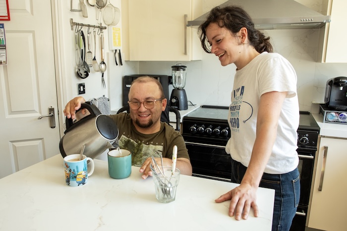 Two people at a table in a kitchen. One in a wheelchair making tea. 