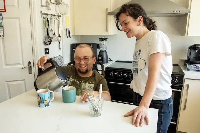 Two people at a table in a kitchen. One in a wheelchair making tea.