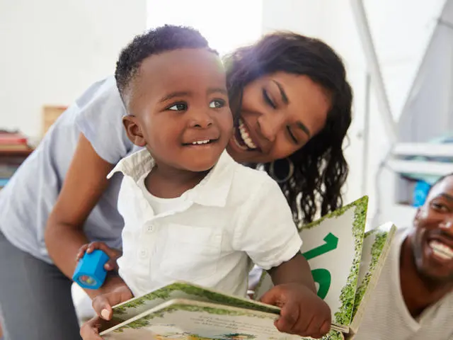 A woman and man are smiling at a small boy. The small boy is holding a book and a blue toy. The small boy is smiling. 