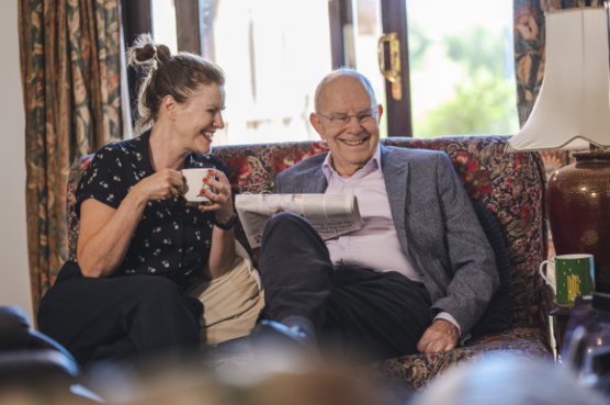 Man sitting with a woman having a cup of tea reading the paper. 