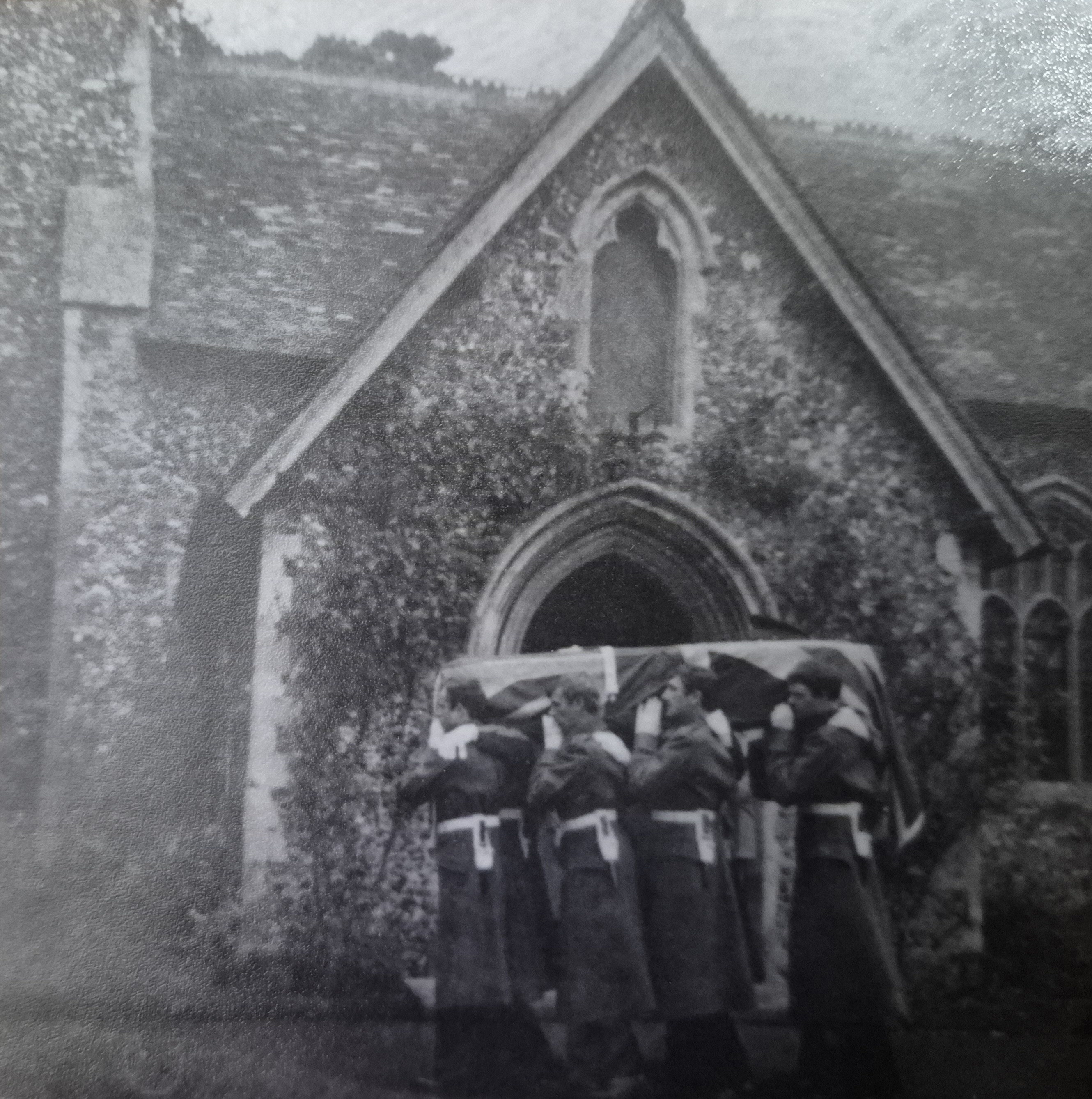 Black and white image of army pall bearers carrying a coffin draped in a union flag outside a church.