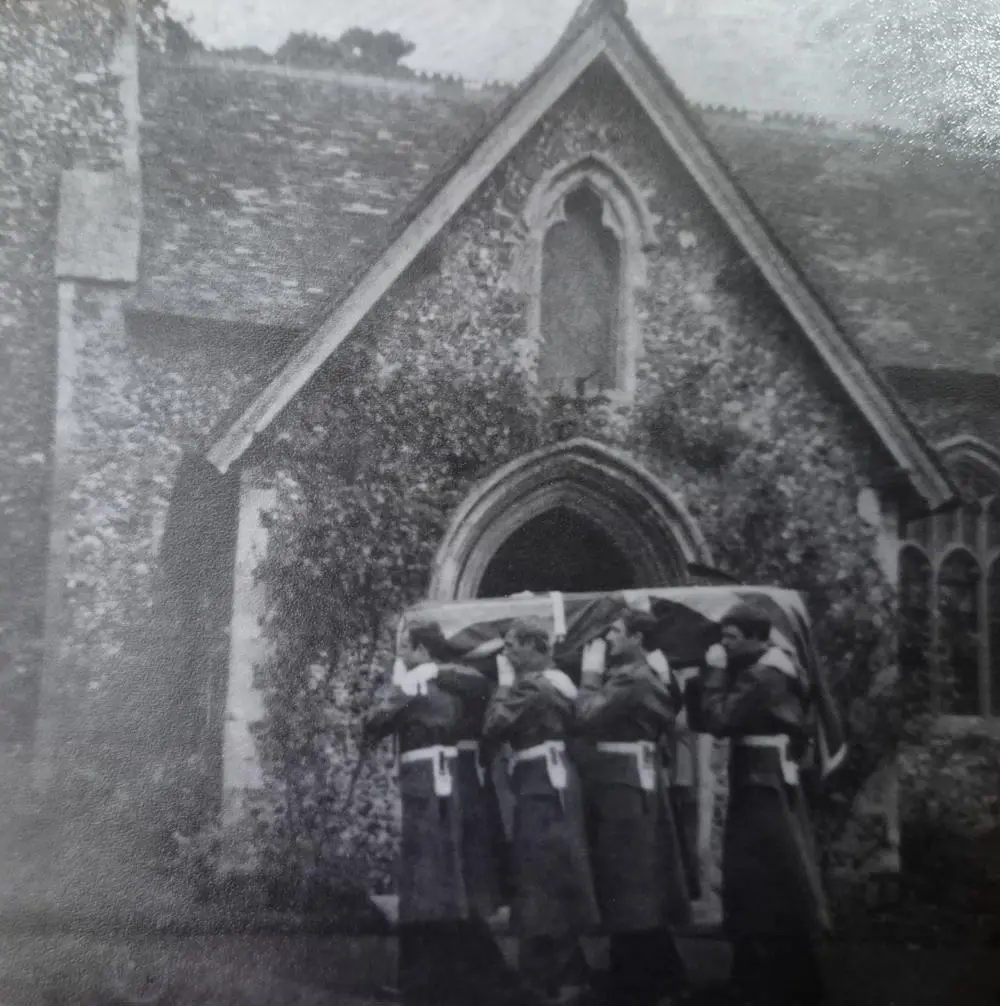 Black and white image of army pall bearers carrying a coffin draped in a union flag outside a church.
