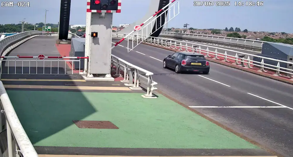 A black car with number plate obscured drives through a red warning light as a barrier comes down.