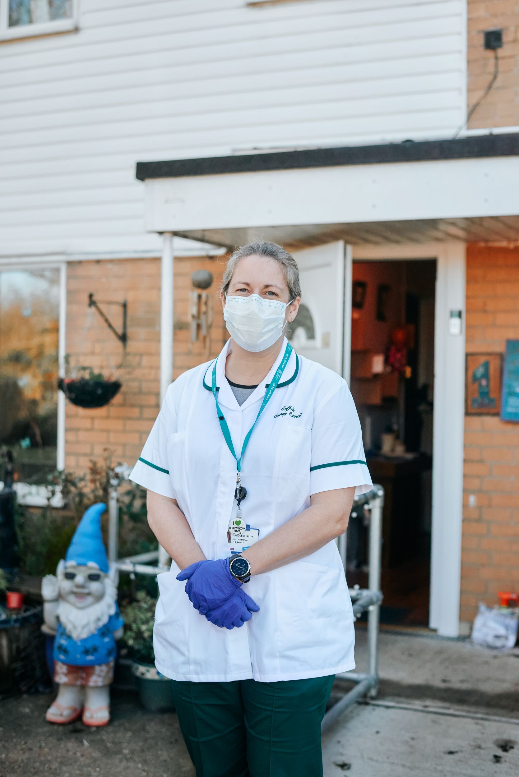 Image of a home first worker standing outside of a house with healthcare uniform on. There is a knome in the garden. 