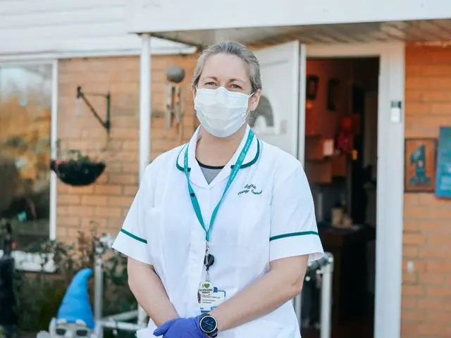 Image of a home first worker standing outside of a house with healthcare uniform on. There is a knome in the garden.