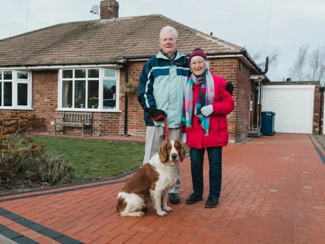 Older couple stand together outside their house with a dog