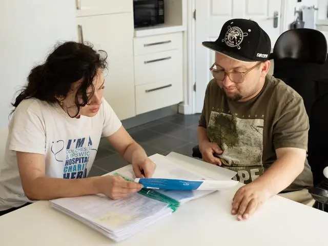 Two people sitting discussing some forms. One person is in a wheel chair and has learning disabilities.