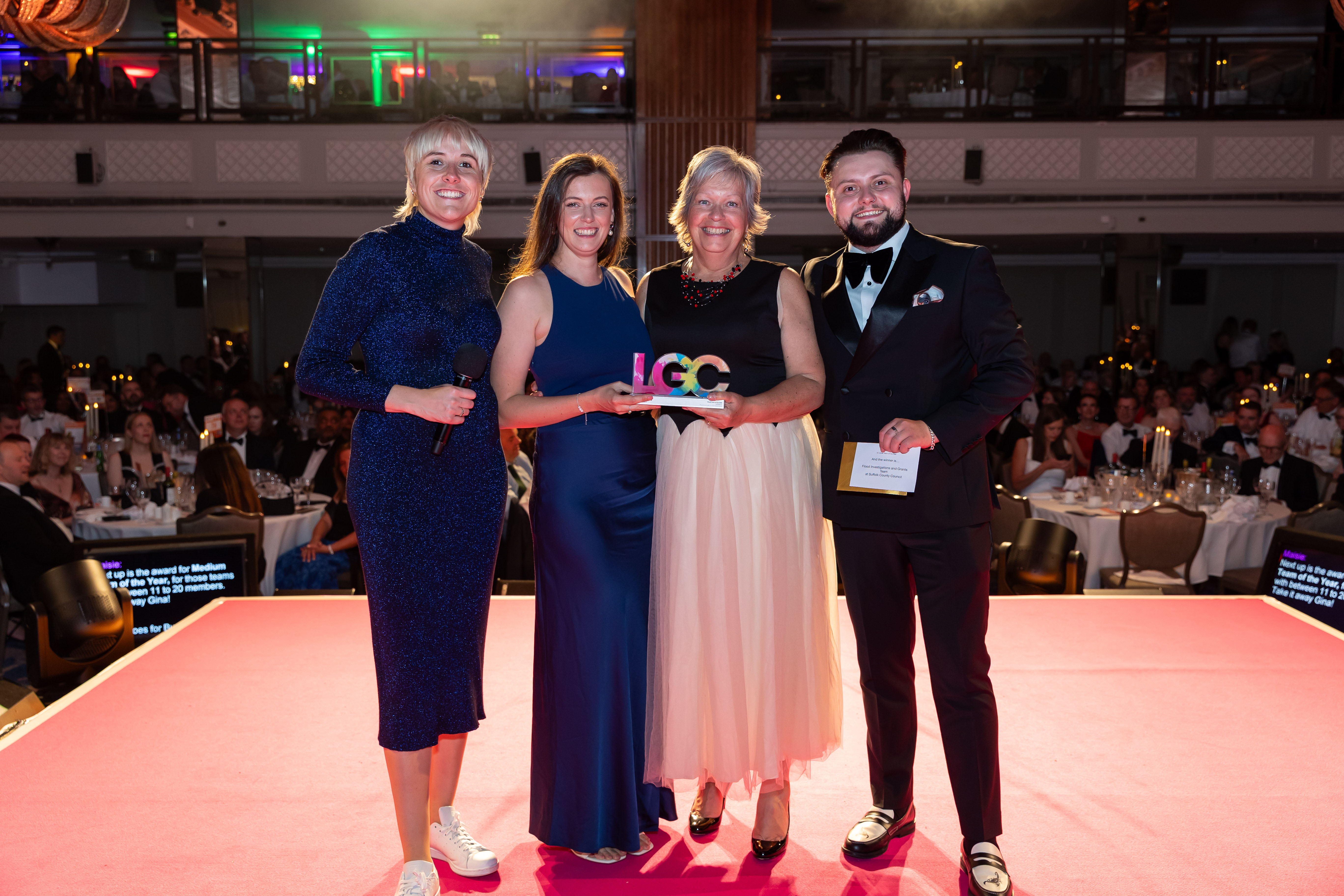 Maisie Adam, Ellie Coleby, Beverly Davies and Kieran McDougall, all in evening dress, on a stage holding the LGC trophy