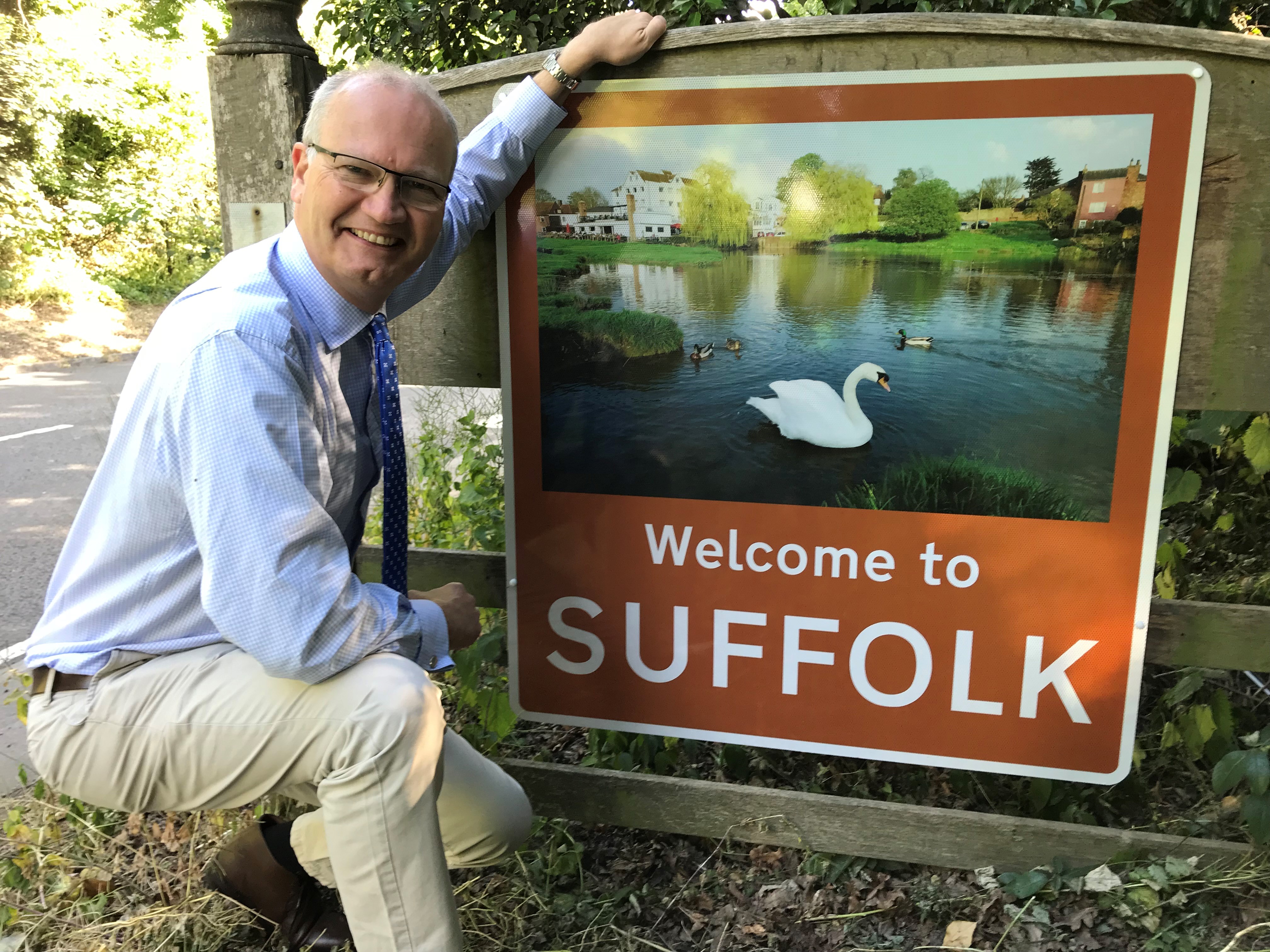 Cllr Matthew Hicks smiling with a brown 'Welcome to Suffolk' sign - it has a photo of a swan in a large pond 