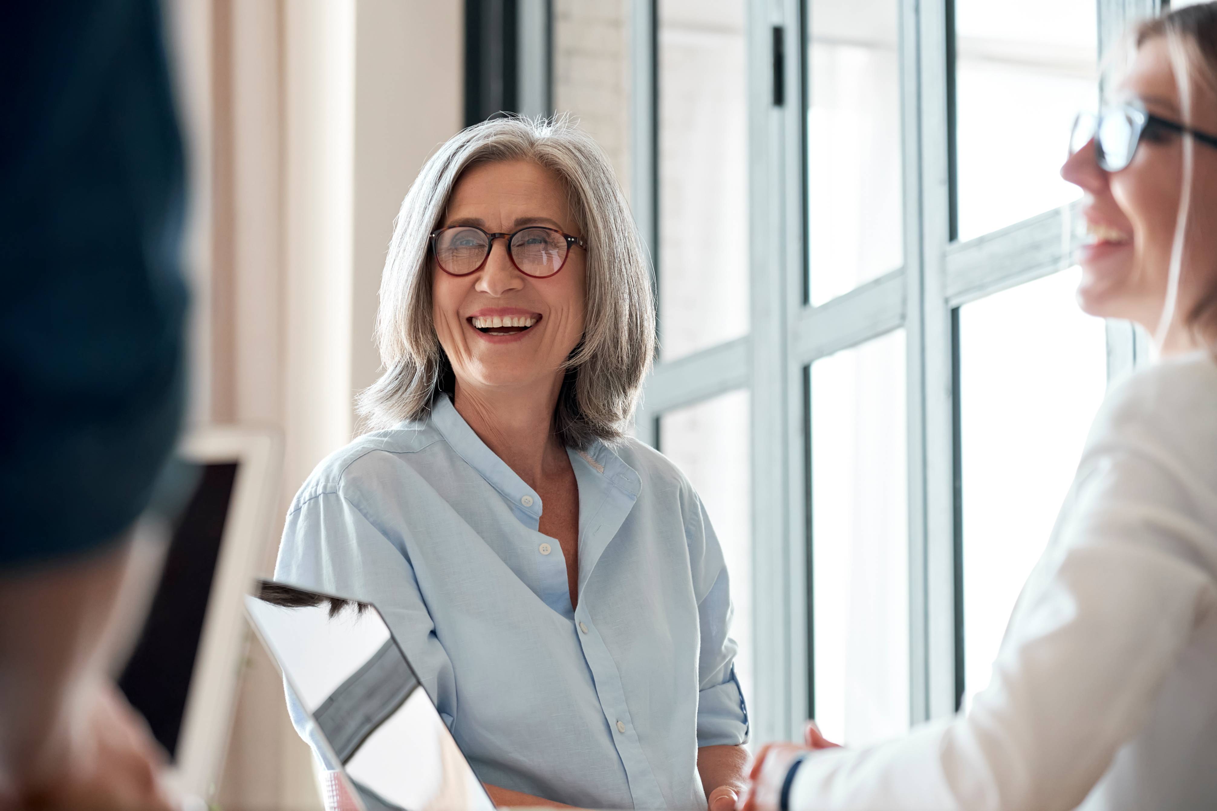 Older mature woman laughing and chatting with colleagues using laptops