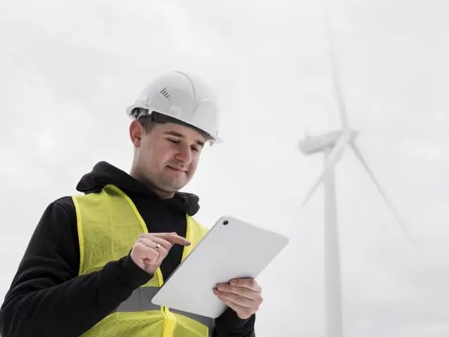Man in hard hat and hi-vis jacket by a wind farm looking at a tablet.