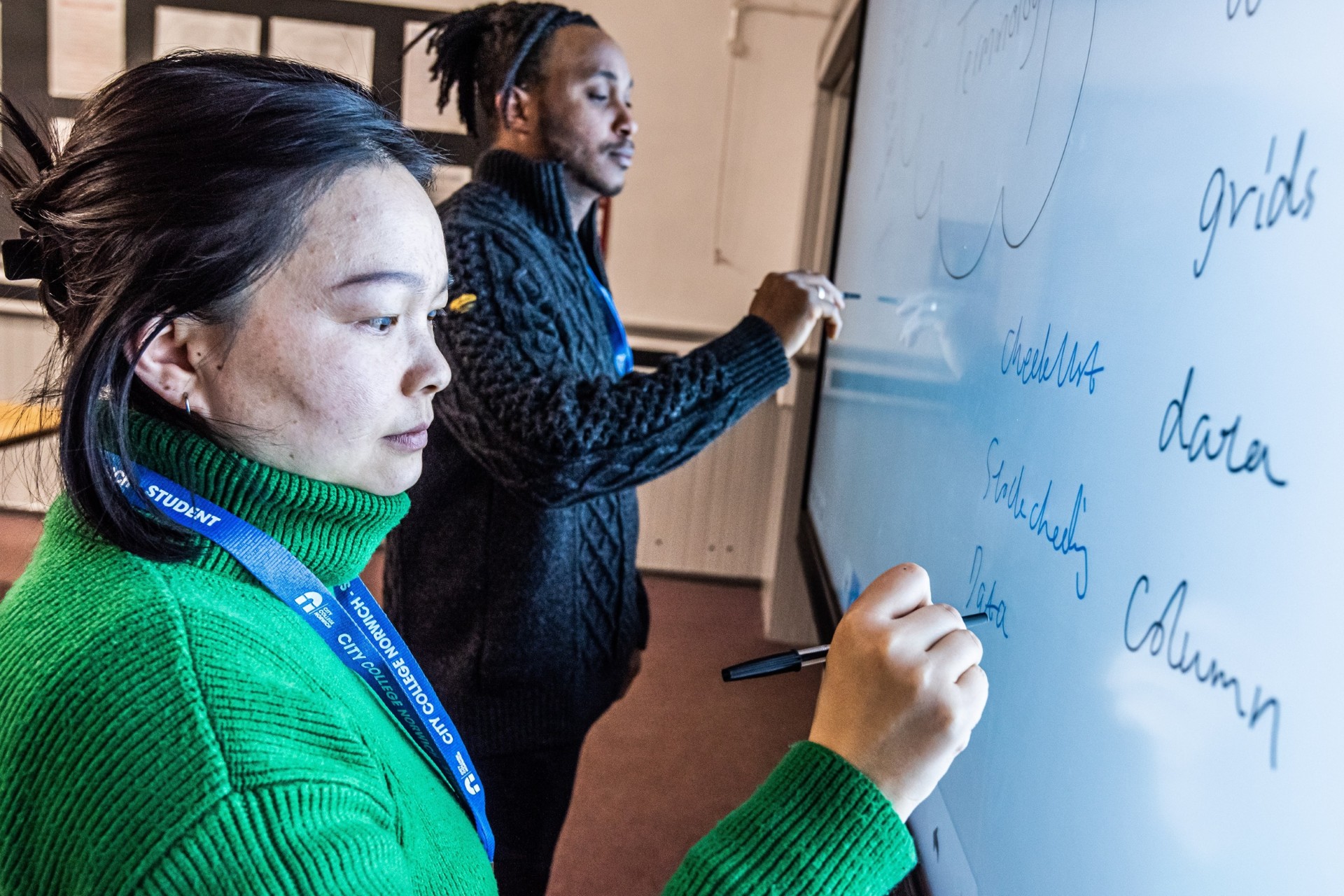 Two people are writing on a whiteboard using blue pens