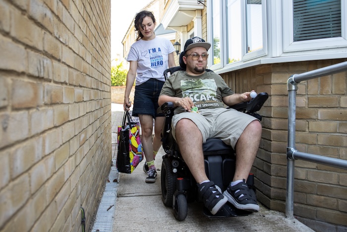 A person in a wheelchair heading home with a charity care worker behind carrying a shopping bag. 