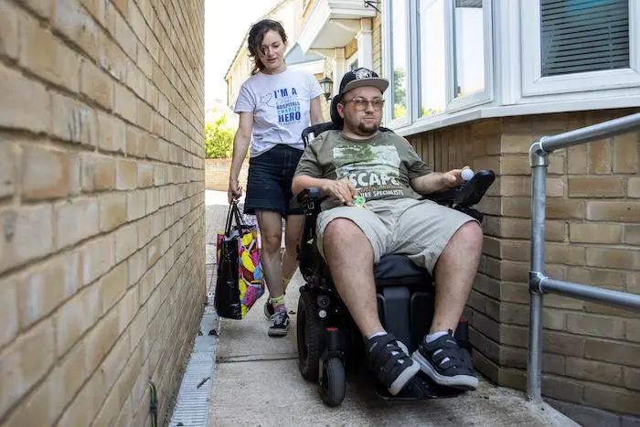 A person in a wheelchair heading home with a charity care worker behind carrying a shopping bag.