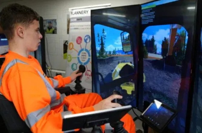 A man in an orange hi-vis jacket is using a simulator of a digger