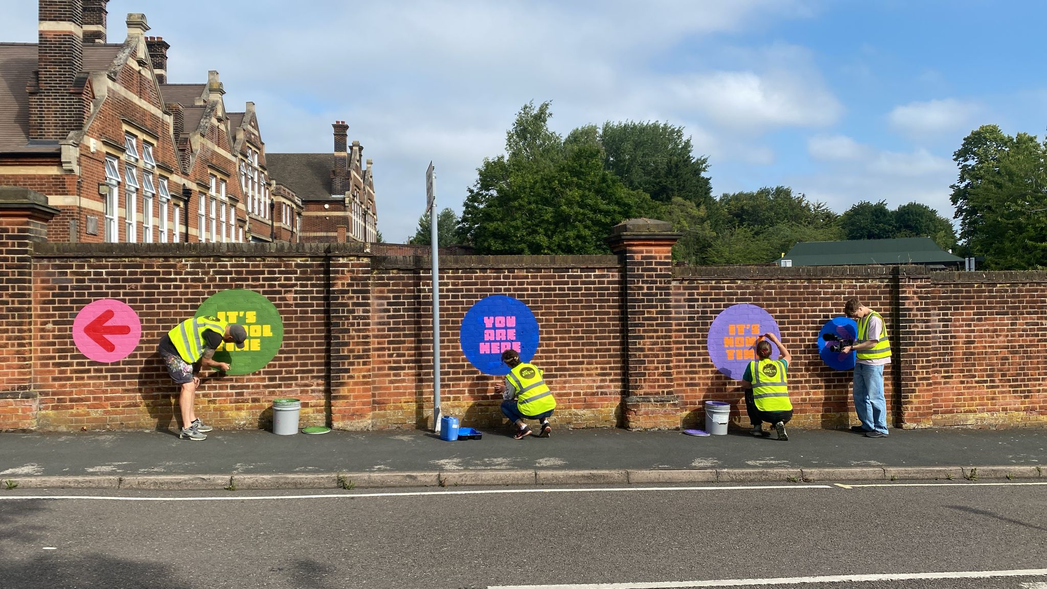 Four people in yellow high viz jackets painting coloured logos on a brick wall.