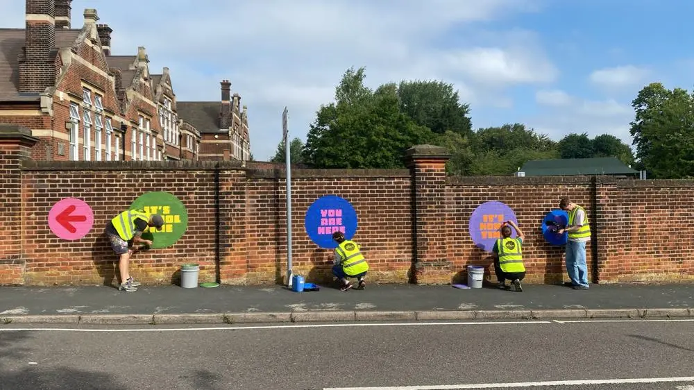 Four people in yellow high viz jackets painting coloured logos on a brick wall.