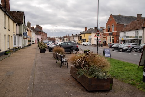 Pavement, verge and road in Long Melford with houses on the left and shops on the right. Plant pots in the middle.