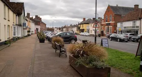 Pavement, verge and road in Long Melford with houses on the left and shops on the right. Plant pots in the middle.