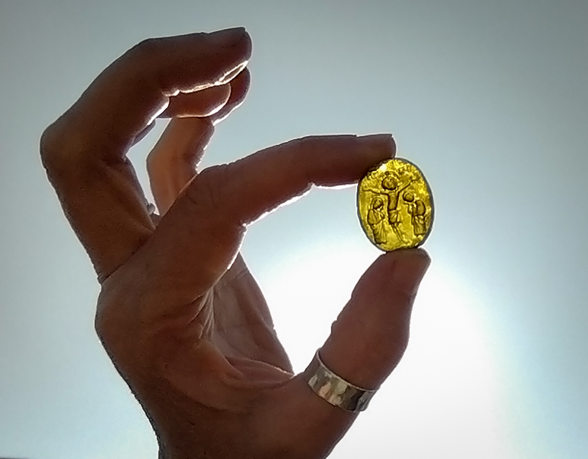 a hand holds the glss ellow glass object up to the light, showing three figures etched into the glass