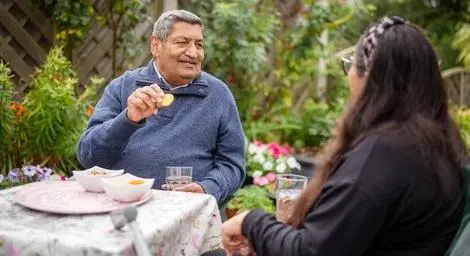 South Asian man over 50 sitting at a table in his garden eating snacks, drinking water, and talking with young woman