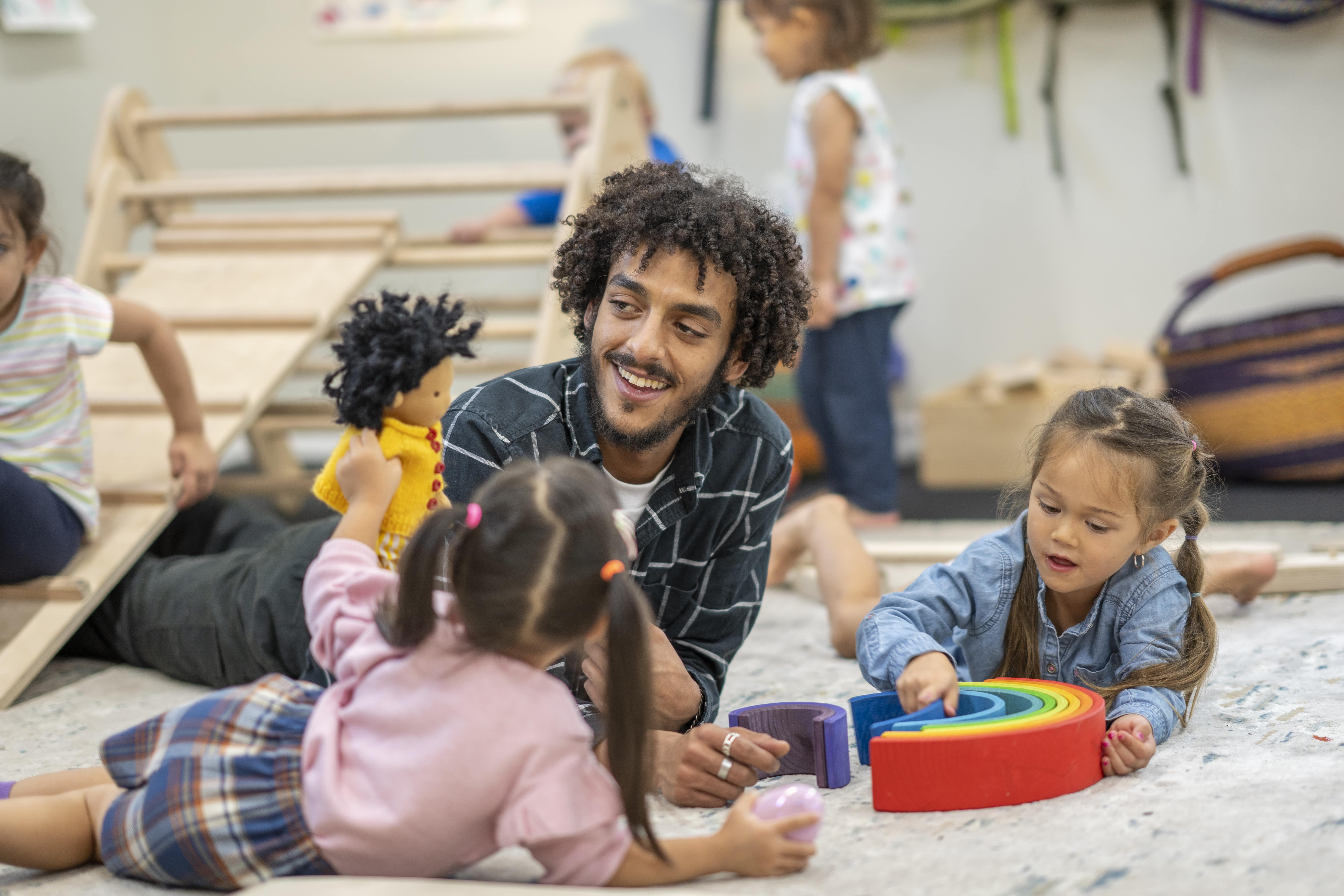 A teacher and young students are laying on the floor, the students are playing with dolls and a rainbow.