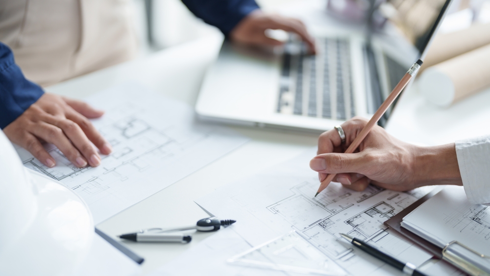 Two people are working over a set of blueprints, one is holding a pencil