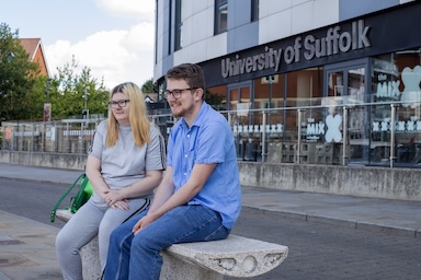 Two people sitting on a bench outside University of Suffolk's Waterfront Building. 