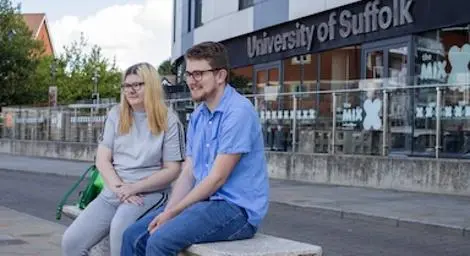 Two people sitting on a bench outside University of Suffolk's Waterfront Building.