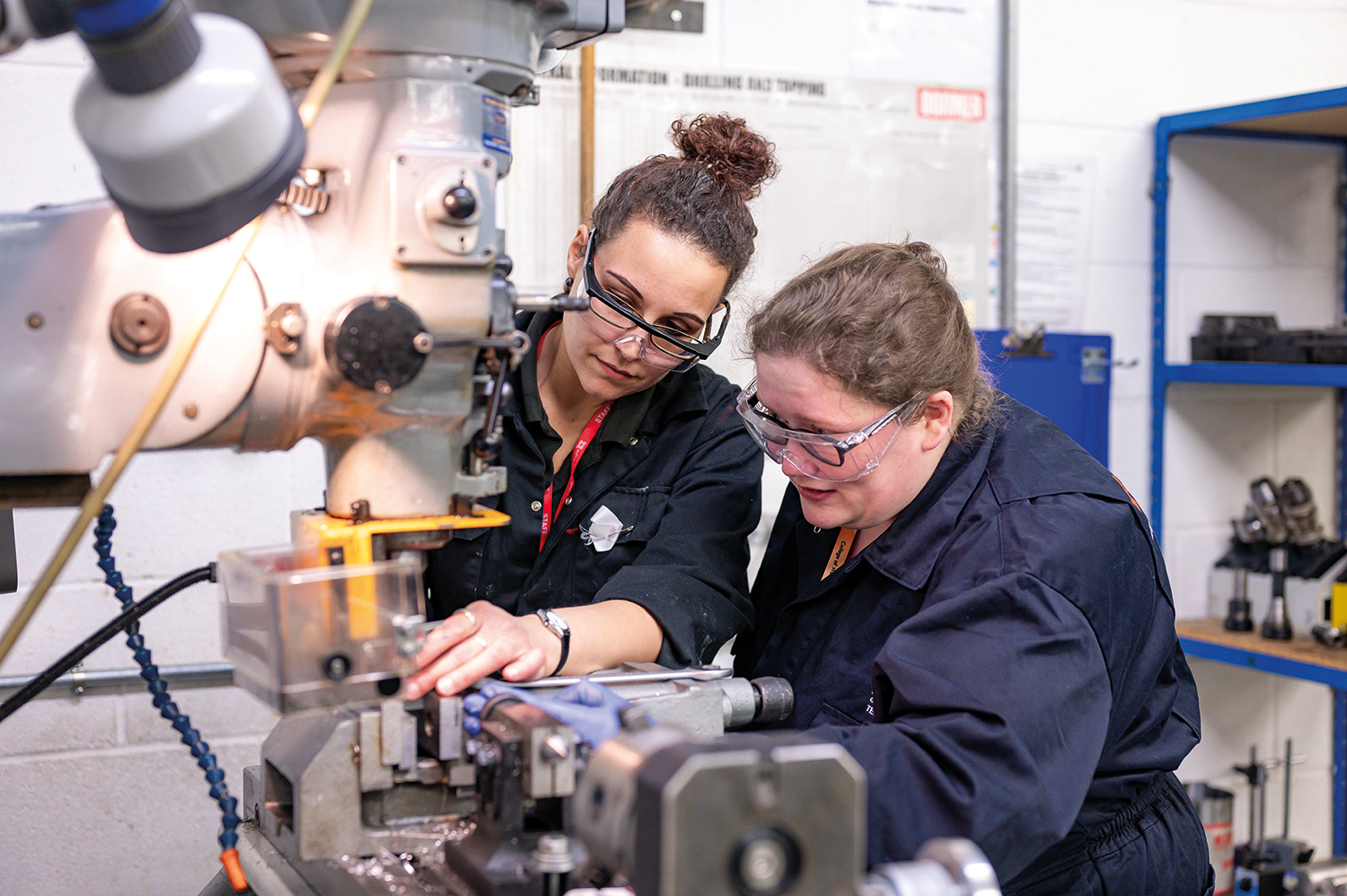 Two women are wearing overalls and wearing protective goggles whilst they operate heavy machinery.