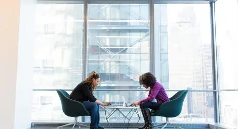 Two women with laptops talk at a table