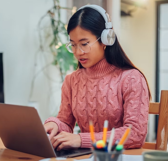 A woman wearing a pink jumper, glasses and headphones, is using a laptop