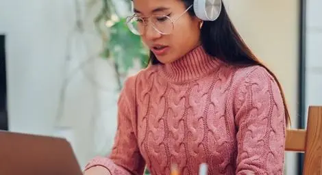 A woman wearing a pink jumper, glasses and headphones, is using a laptop